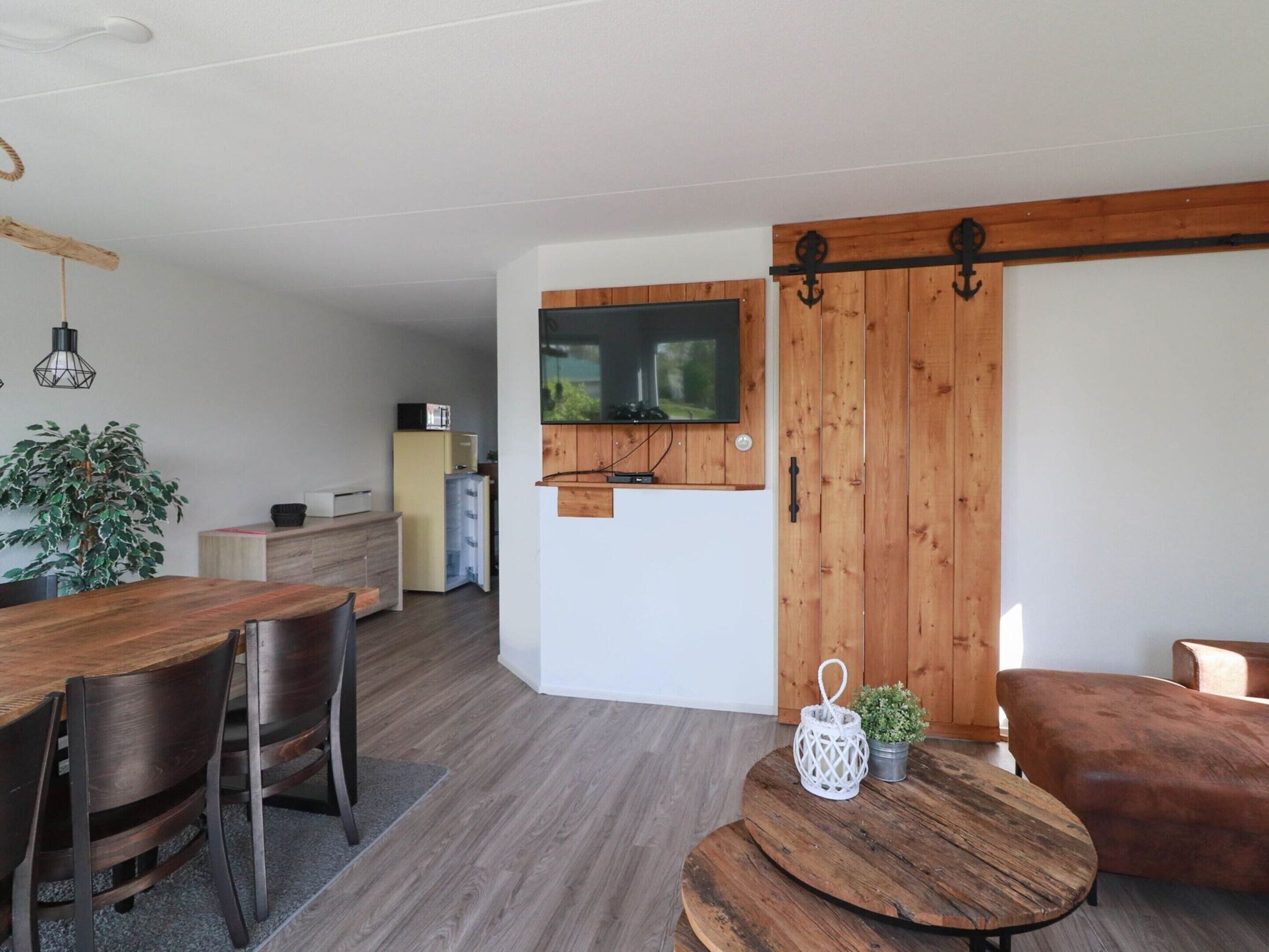 A modern living room with a wooden dining table, chairs, a TV mounted on a wooden wall, a sofa, and a small round table with a plant.