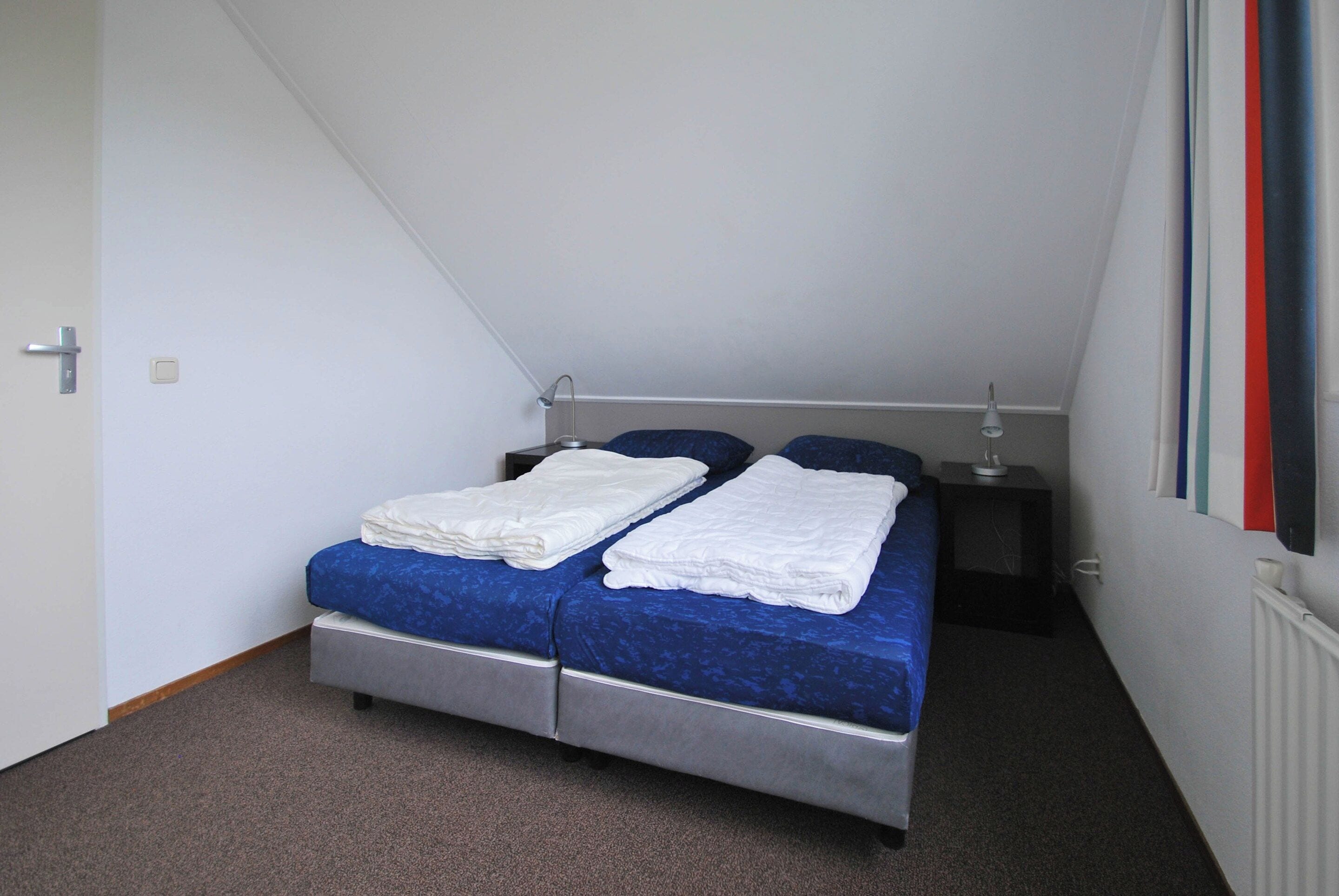A small, single-bed room with a sloped ceiling, a bedside table with a lamp, and a radiator.