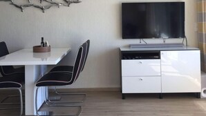 A modern dining area with a white table, black chairs, a flat-screen TV, and a white cabinet with a dishwasher. - Apartment Holiday Harbor (Grossenbrode)