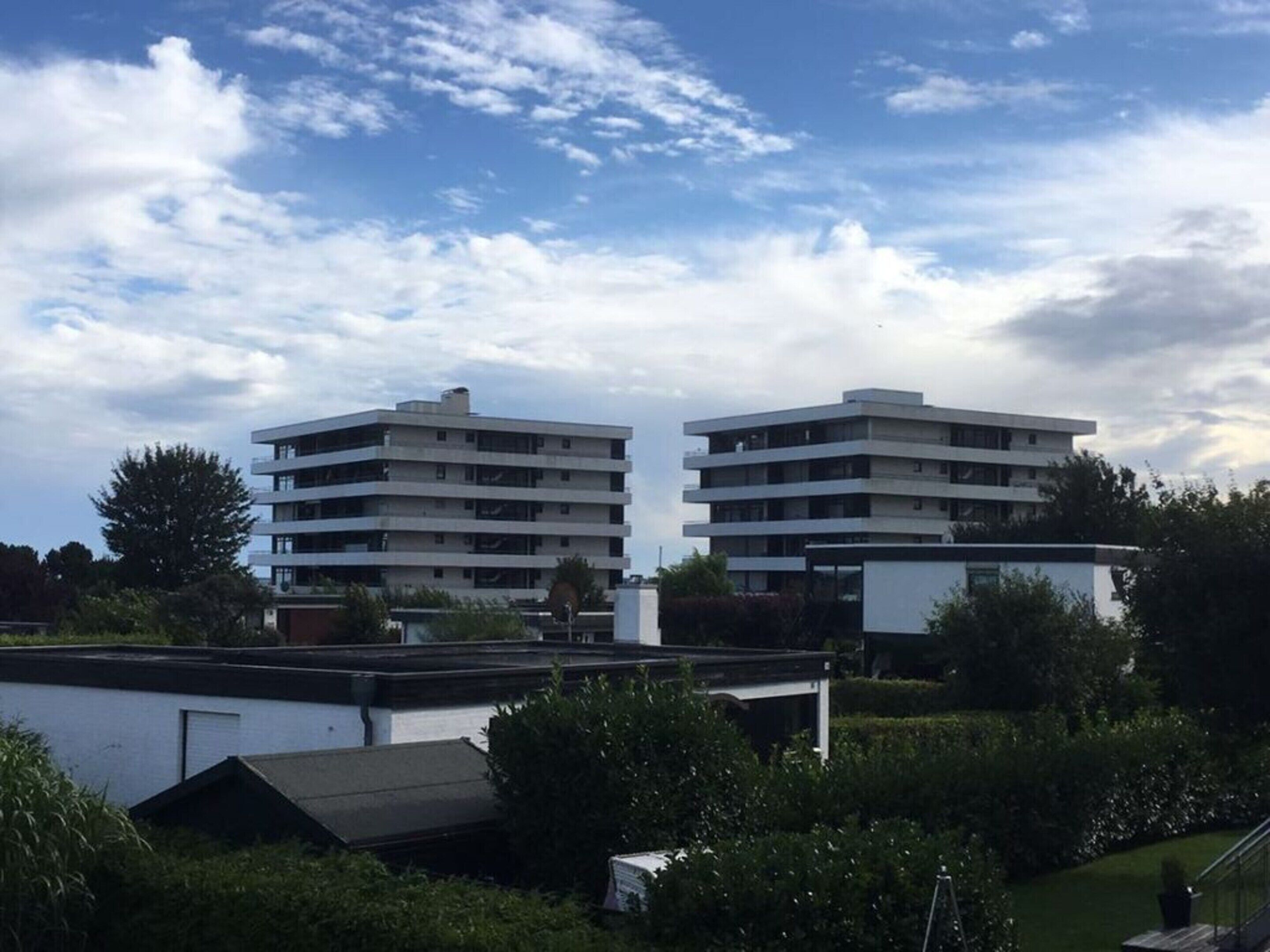 Deux bâtiments modernes à plusieurs étages, dotés de toits plats, entourés de verdure et baignés d’un ciel dégagé.
