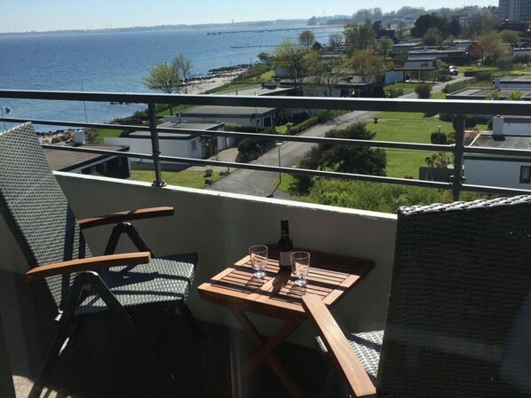 Un balcon avec vue sur la mer et un pont, une table avec une bouteille et des verres, et une chaise.