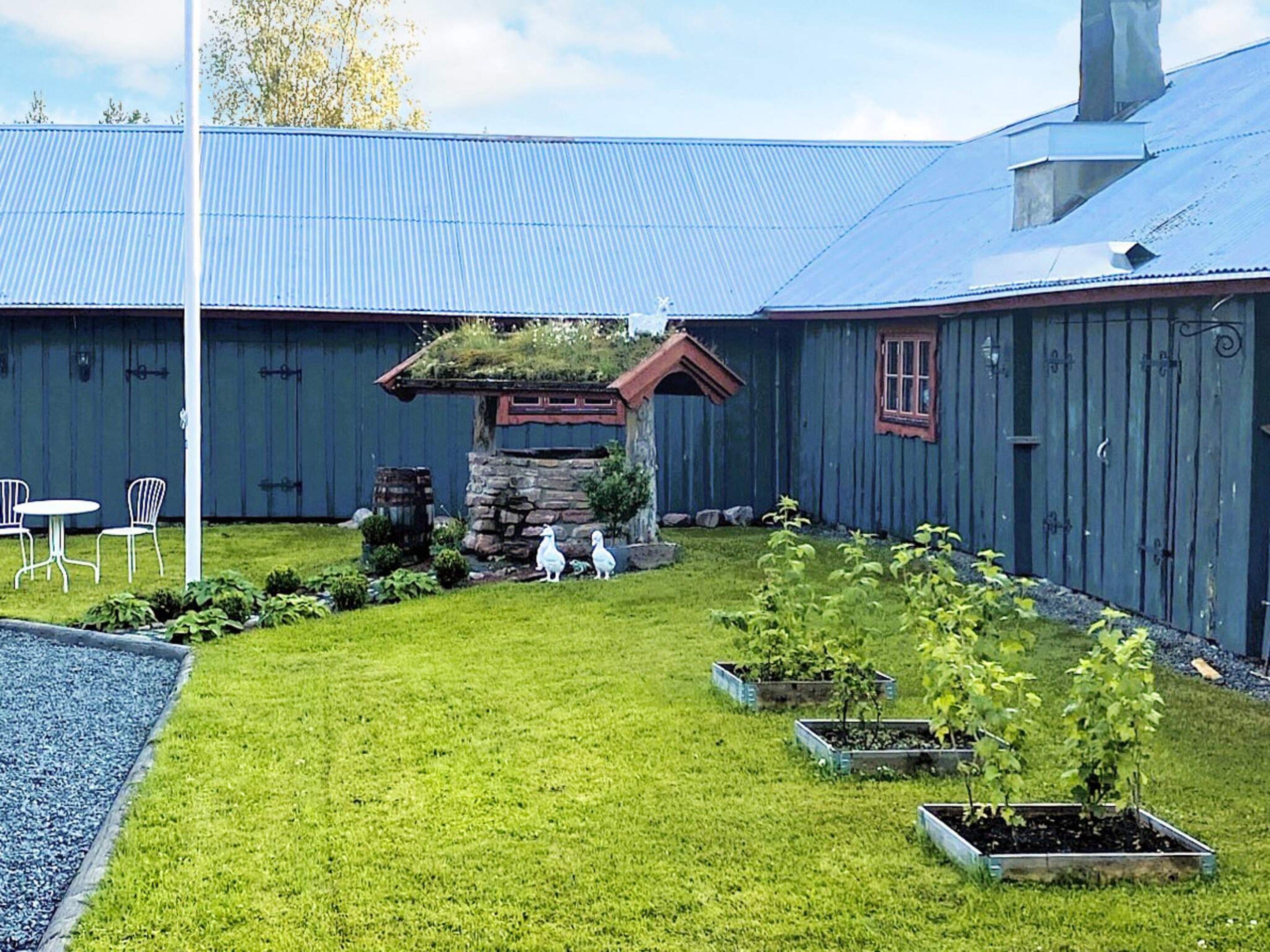 A building with a blue roof, a stone structure with a red roof, and a grassy area with plants and two white chairs.