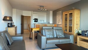 A modern living room with a grey sofa, a small coffee table, and a dining area with wicker chairs. - Apartment in Nieuwpoort With sea View (Nieuwpoort)