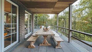 Une terrasse extérieure avec une table et des bancs en bois, entourée de baies vitrées et d’une balustrade donnant sur une zone boisée.