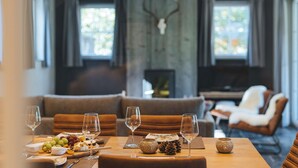 Un coin repas avec une table en bois dressée pour un repas, où l’on trouve une variété de fromages, de fruits et de noix. Des verres à vin sont posés sur la table, et l’ensemble comprend des sièges confortables et une grande poutre en bois ornée de bois de cerf en arrière-plan.