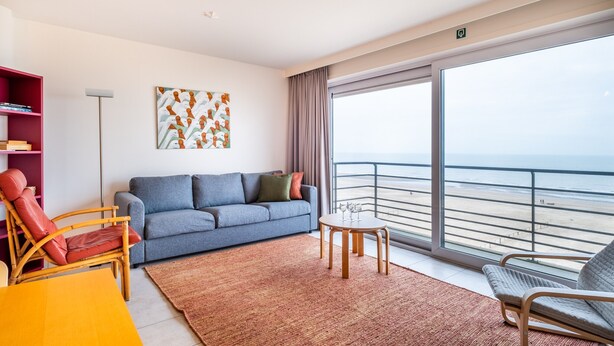 Ein modernes Wohnzimmer mit einem grauen Sofa, einem roten Sessel, einem kleinen runden Tisch mit Gläsern und einem großen Fenster mit Blick auf einen Strand.