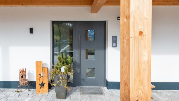 A modern building entrance with a dark grey door, wooden beams, a metal bench, and a potted plant with greenery. - Apartment in Uderns Near Spieljoch Ski (Uderns)