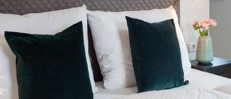 Una cama con almohadas blancas y verde oscuro, un cabecero acolchado gris y un florero con flores sobre una mesita de noche.
