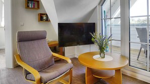 Ein modernes Wohnzimmer mit großem Fenster, einem Flachbildfernseher, einem bequemen Sessel, einem kleinen Holztisch und einem Bücherregal.