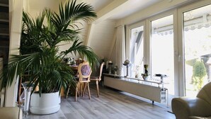 Une pièce avec une grande plante en pot, une table à manger avec des chaises et une fenêtre donnant sur l’extérieur.
