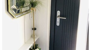 Une porte sombre avec une poignée argentée, un miroir aux motifs géométriques accroché au mur, et une étagère blanche ornée d’une plante et d’une fleur blanche.