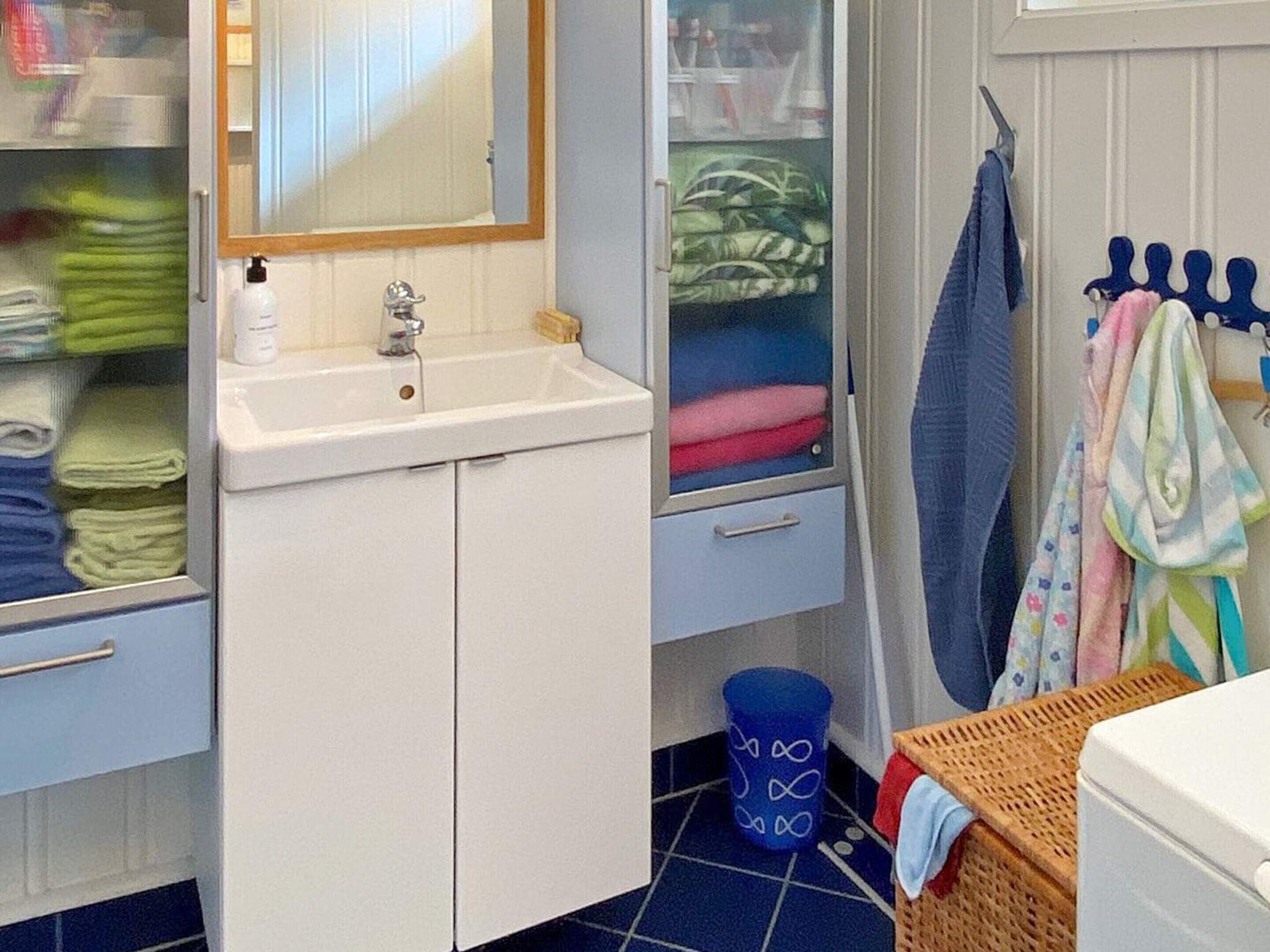 Ein Badezimmer mit Waschbecken, Spiegel und Stauraumschränken. Ein blauer Mülleimer und ein Korb stehen auf dem Boden.