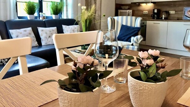 Ein Essbereich mit einem Holztisch, Stühlen und Zimmerpflanzen. Im Hintergrund befindet sich eine Couch und eine Küche.