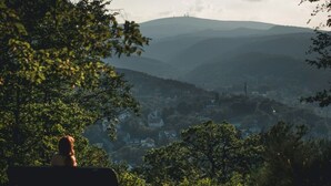 Eine Person sitzt auf einer Bank und blickt auf eine Stadt mit Hügeln im Hintergrund.