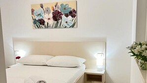 A neatly made bed with white linens and pillows, a bedside table with a lamp, and a floral painting above the bed. - Apartments Jelena - One Bedroom Apartment (Dubrovnik)