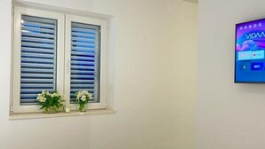 A room with a window with blinds, a TV mounted on the wall, and a vase with flowers on the windowsill. - Apartments Jelena - One Bedroom Apartment (Dubrovnik)