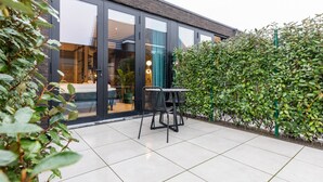Un bâtiment moderne doté de grandes fenêtres en verre, d’un patio avec une table et des chaises, et d’une haie verdoyante.