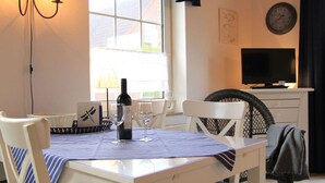 A dining area with a table set for two, a bottle of wine, and two wine glasses. There is a television on a cabinet, a window with a view of the outside, and a chair with a blanket. - Overt Meer 6 Modern Retreat (Grosefehn)