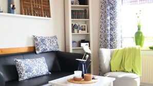 A living room with a black sofa, patterned cushions, a white coffee table, a bookshelf, and a window with curtains. - Overt Meer 6 Modern Retreat (Grosefehn)