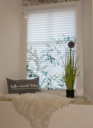 Une fenêtre donnant sur des bambous, un coussin décoratif orné d’un motif textuel et une plante en pot posée sur un rebord de fenêtre.