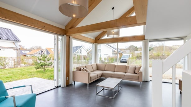 A modern living room with a large L-shaped beige sofa, a small coffee table, and a blue armchair. The room features a high ceiling with exposed wooden beams and large windows offering a view of the outside. - Holiday Home in Renesse Near the Beach (Renesse)