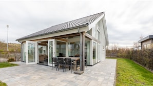 A modern house with a covered patio, a dining area with chairs, and a large window. - Holiday Home in Renesse Near the Beach (Renesse)