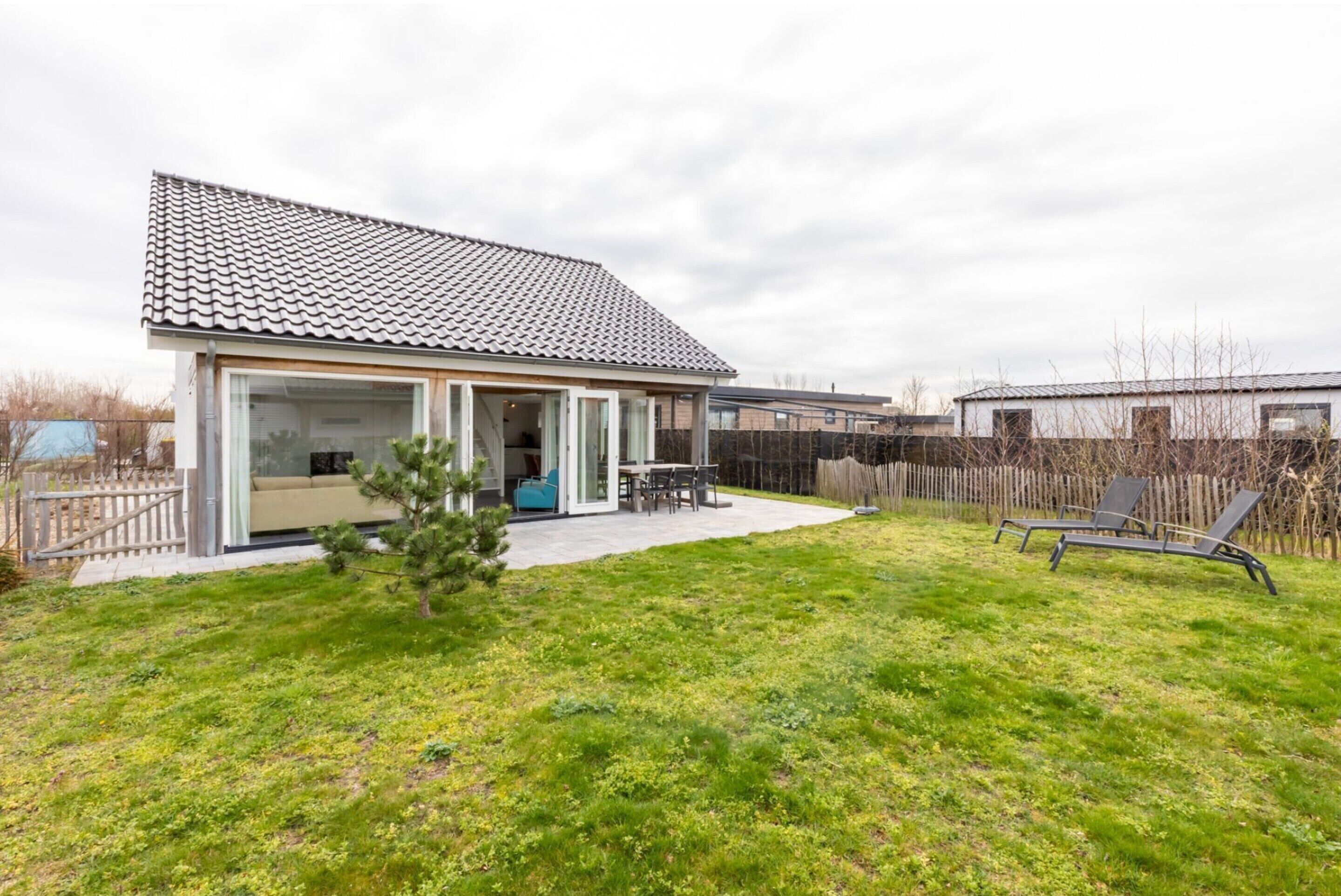 A modern house with a tiled roof, a glass door entrance, a patio with a table and chairs, and a fenced backyard.