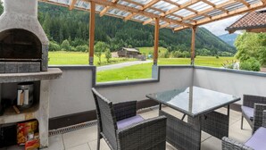 Un patio techado con un horno de piedra, mobiliario de mimbre y vistas a un paisaje verde con montañas.