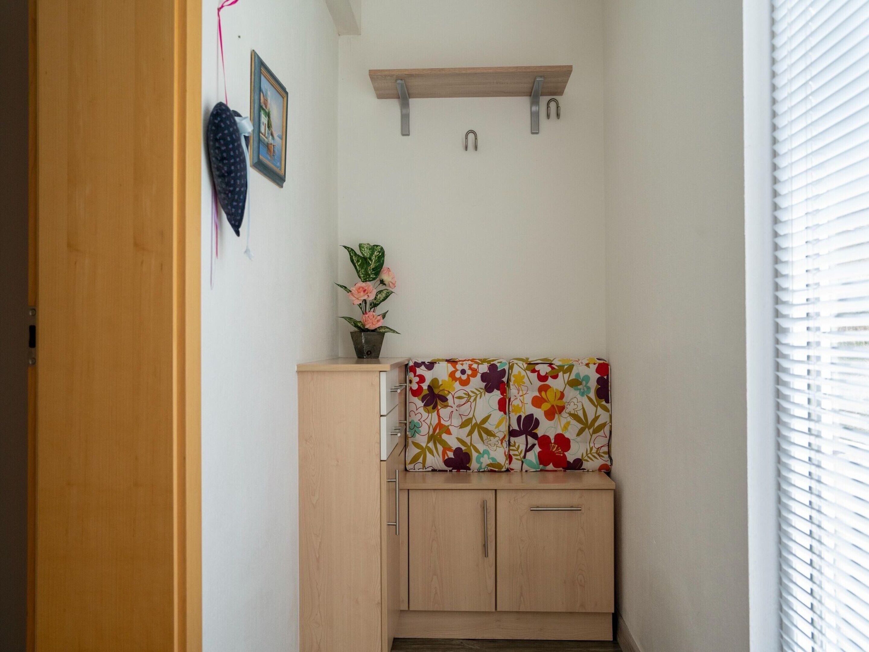 A small, neatly organized room with a wooden cabinet, a floral-patterned cushion, a small potted plant, and a picture frame on the wall.