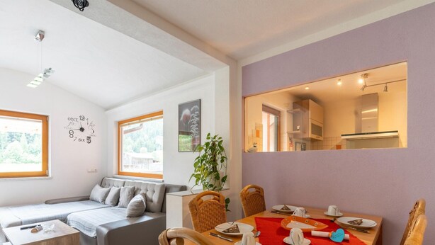 Una sala moderna con sofá, mesa de comedor y sillas. Se ve un ventanal grande, una planta y una cocina a través de una puerta abierta.