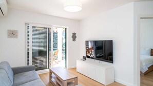 A modern living room with a grey sofa, a wooden coffee table, a TV on a white cabinet, and a sliding glass door leading to an outdoor area. - Apartment Villa Claudia (Dubrovnik)