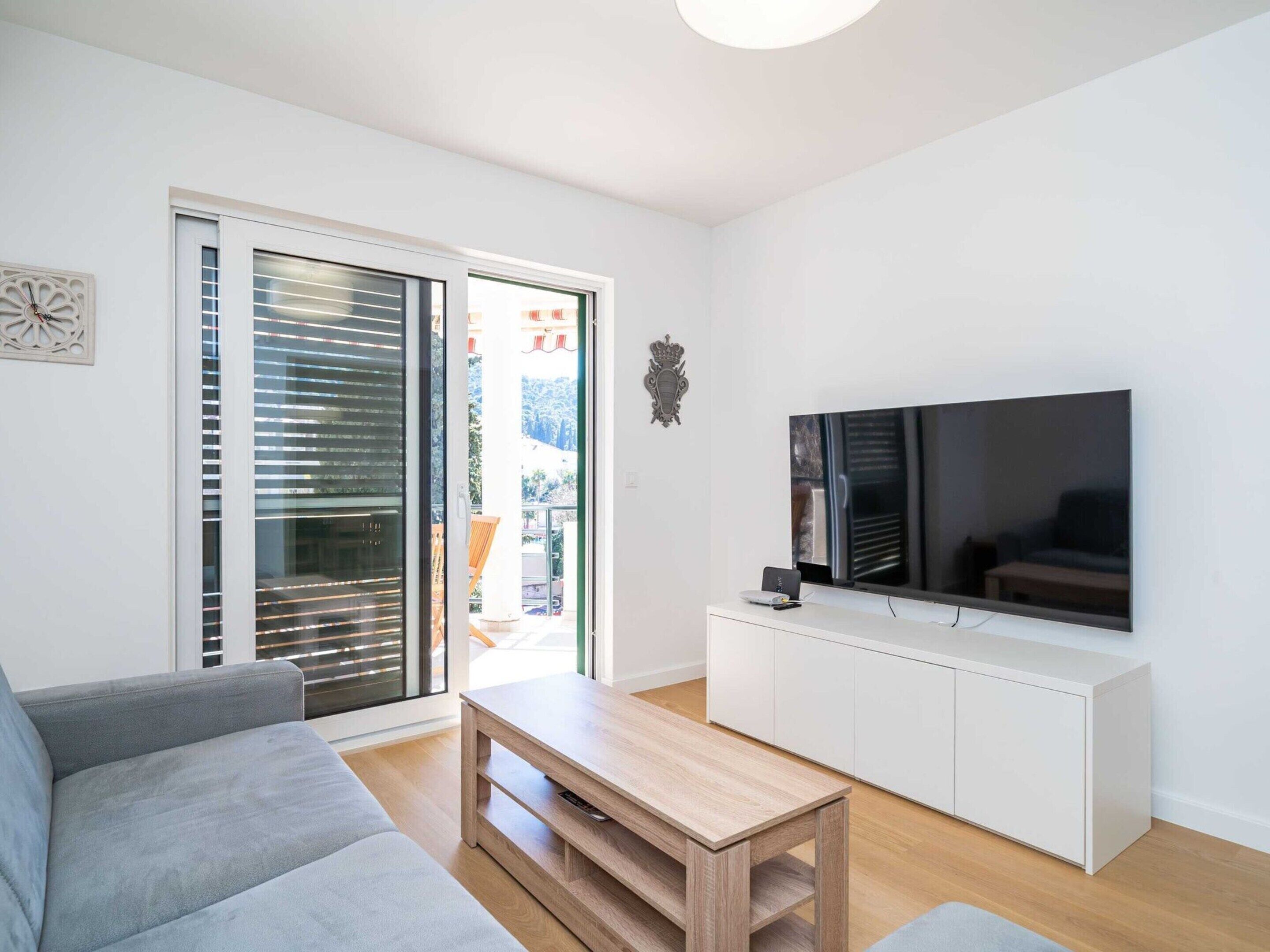 Ett modernt vardagsrum med en grå soffa, ett soffbord i trä, en tv på ett vitt skåp och en skjutdörr i glas som leder ut till ett utomhusområde.