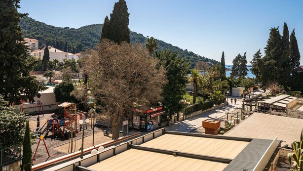 A rooftop terrace with a view of a playground, a building with a red sign, and a hilly landscape in the background. - Apartment Villa Claudia (Dubrovnik)