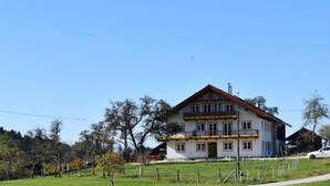 Una casa de dos pisos con un balcón, rodeada por un patio cercado y árboles.