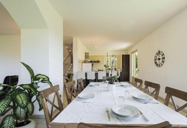 Un comedor con una larga mesa dispuesta para seis personas, rodeada de sillas de madera. Hay plantas en macetas a la izquierda.