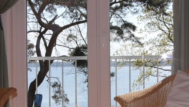 A room with a view of a lake and trees, a wicker chair, and a decorative bird sculpture. - Holiday Home Water View (Rankwitz)