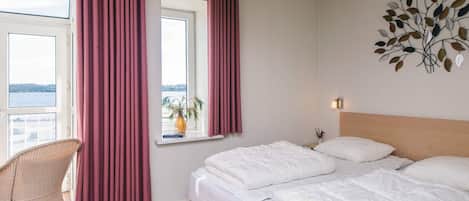 Una camera d'albergo con un letto grande, una finestra con tende, vista sull'acqua, una sedia e un elemento decorativo a parete.