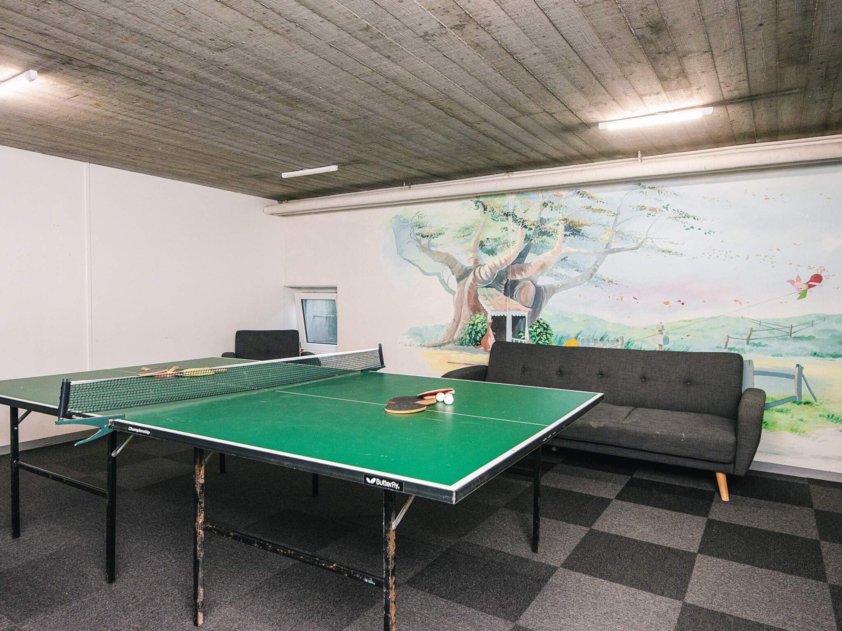 Une table de ping-pong avec un filet, un canapé et une fresque représentant un arbre et un paysage sur le mur.