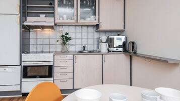 Cocina pequeña con estufa, horno y cafetera. Hay una mesa de comedor con tazas blancas y una silla.