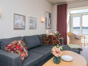 A living room with a sofa, floral cushions, a vase with flowers, and a view of a body of water through a window. - 4 Person Holiday Park Home in Hejls (Hejls)