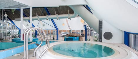 An indoor swimming pool with a hot tub, surrounded by blue and white walls, and a covered seating area with tables and chairs.