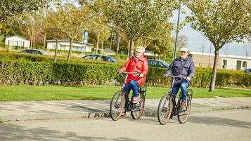 Two people riding bicycles on a paved path in a park-like setting with greenery and buildings in the background.
