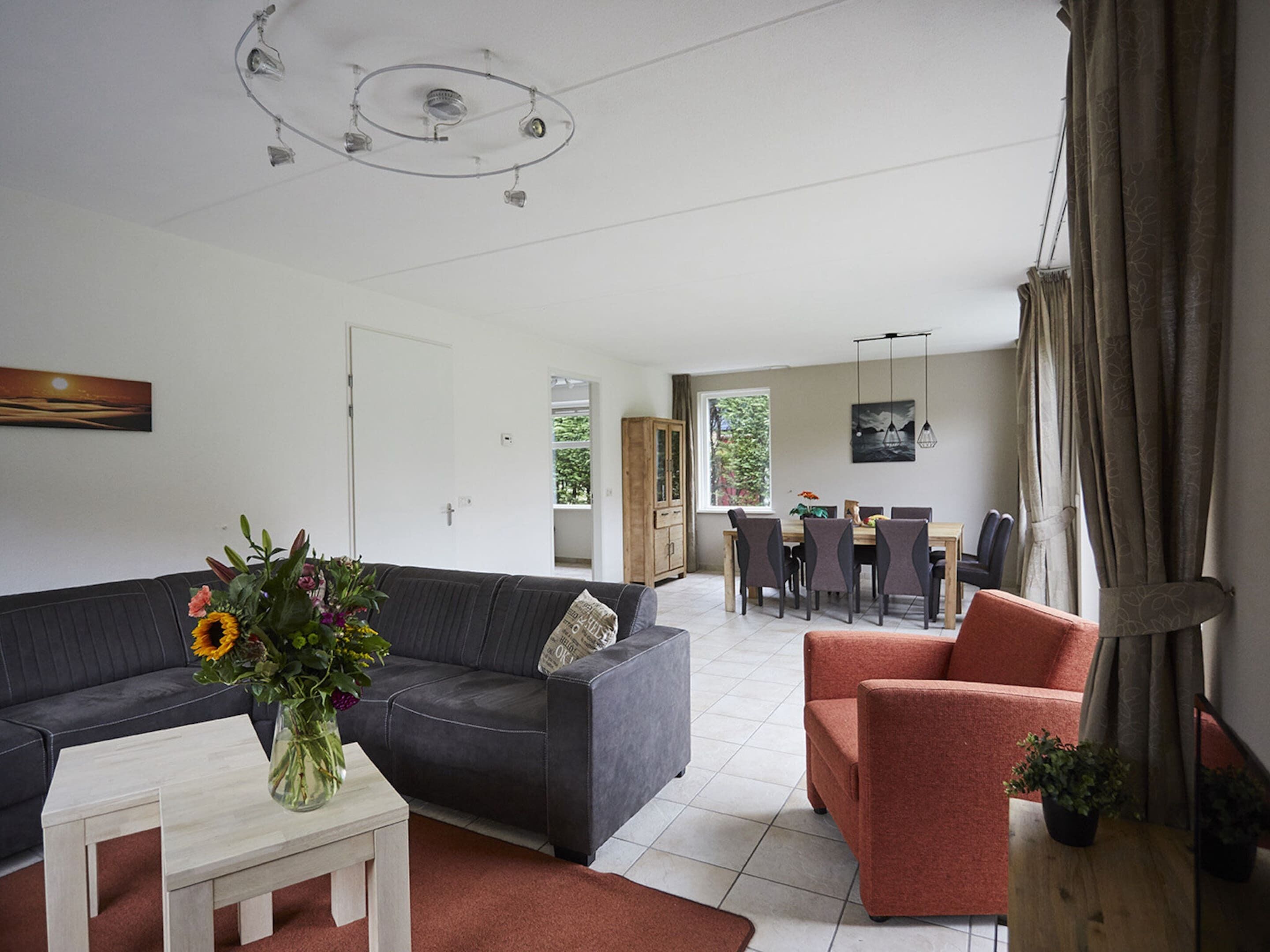 A modern living room with a grey sofa, a red armchair, and a white coffee table. There is a dining area with a table and chairs visible through an open door.