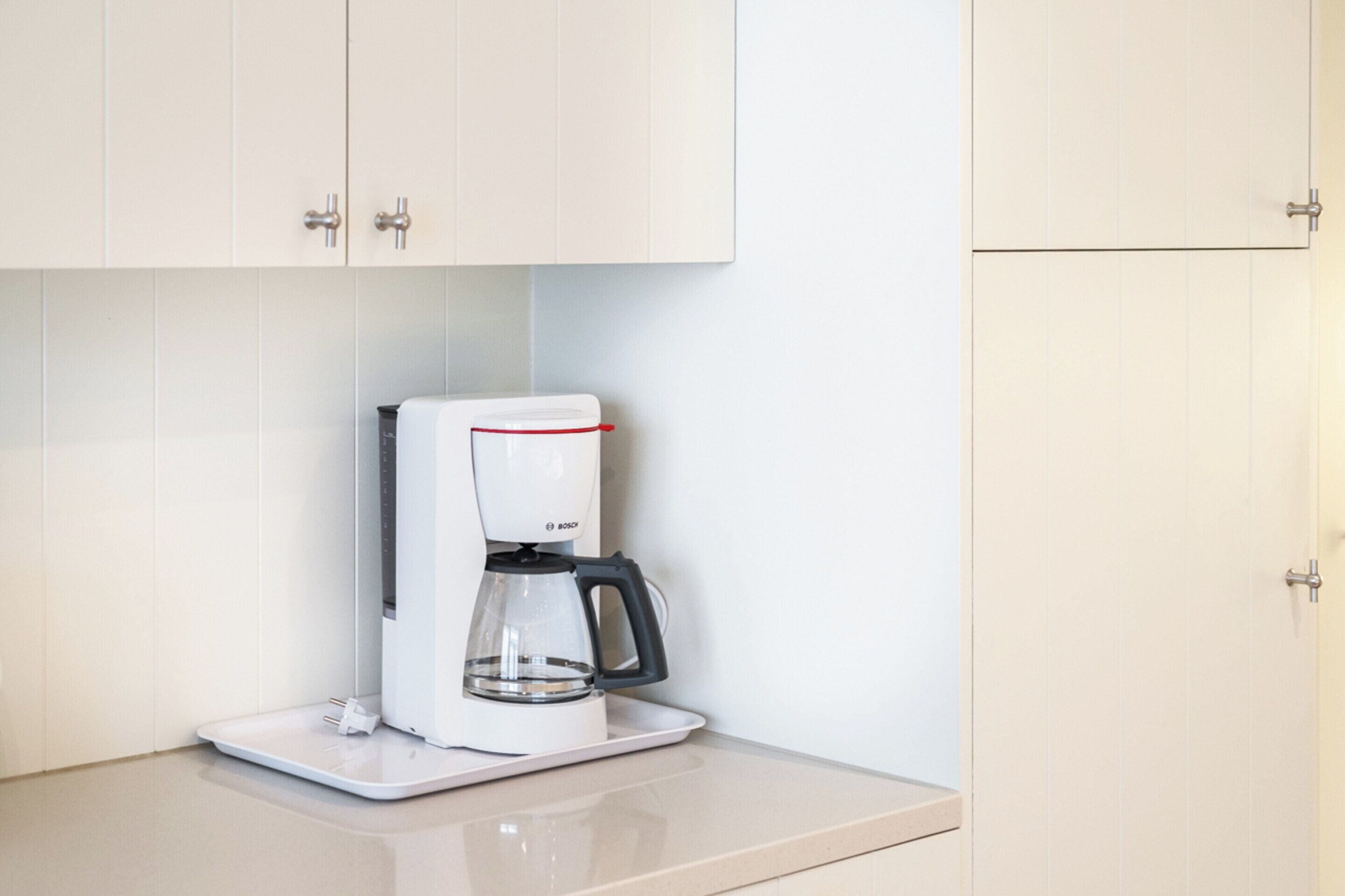 A white coffee maker on a white countertop in a kitchen with white cabinets.