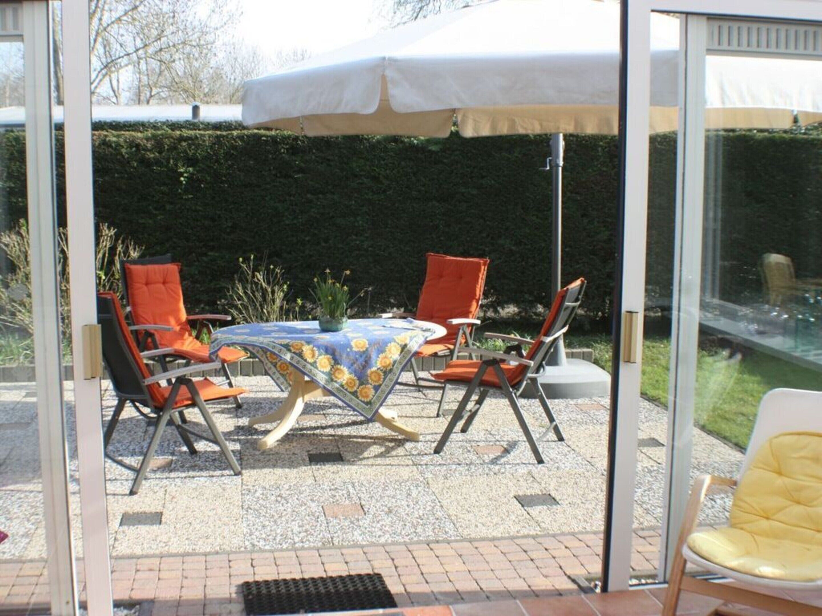 A glass-enclosed patio with a table and four chairs, a white umbrella, and a potted plant.