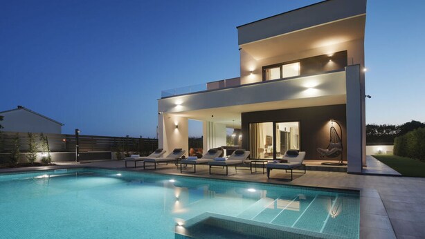 Een modern huis met twee verdiepingen, een zwembad, buitenmeubilair en een goed verlicht terras.
