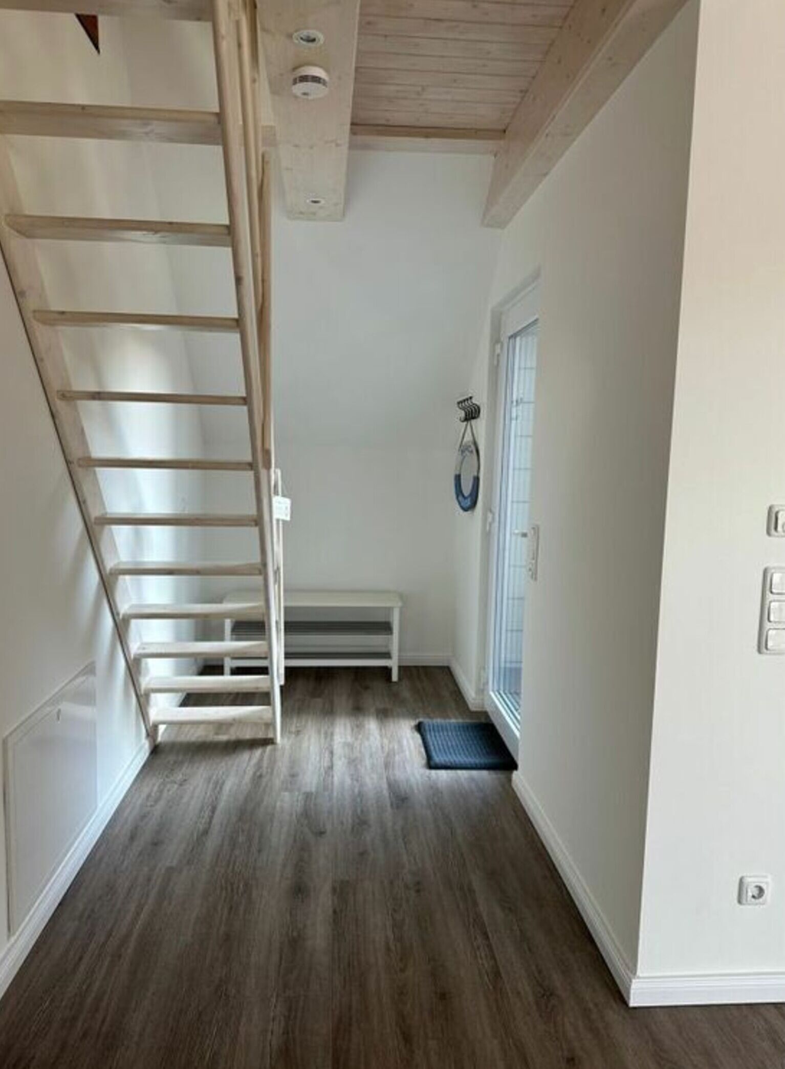 A narrow hallway with a wooden staircase, a bench, and a door with a window.