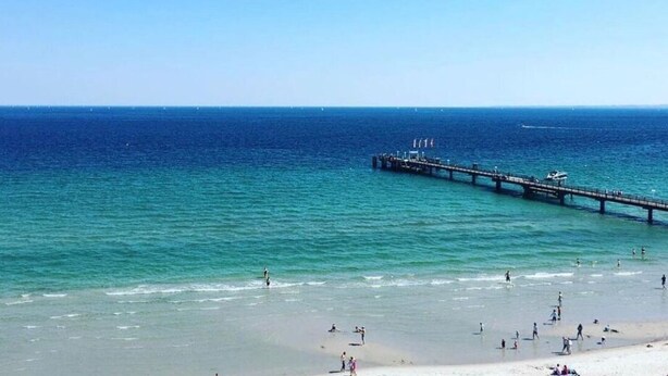 A beach with a pier extending into the ocean, people on the shore, and a clear sky. - Holiday Vacation by the sea (Scharbeutz)