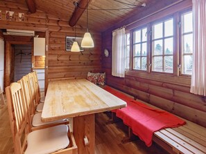 A wooden cabin interior with a dining table, chairs, a bench, a hanging light fixture, and a window with curtains.