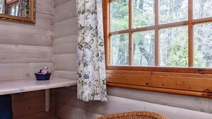 A cabin interior with a wooden wall, a window with a view of trees, a wicker chair, a small table with a blue cloth, and a curtain with a floral pattern. - 6 Person Holiday Home in Vaeggerlose (Vaeggerlose)